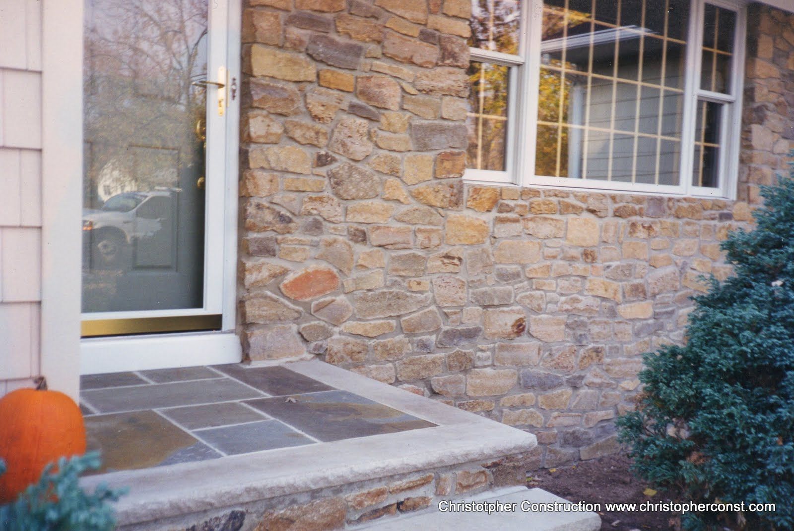 Stone house entrance with slate steps, white door, pumpkin, and shrub beside a window