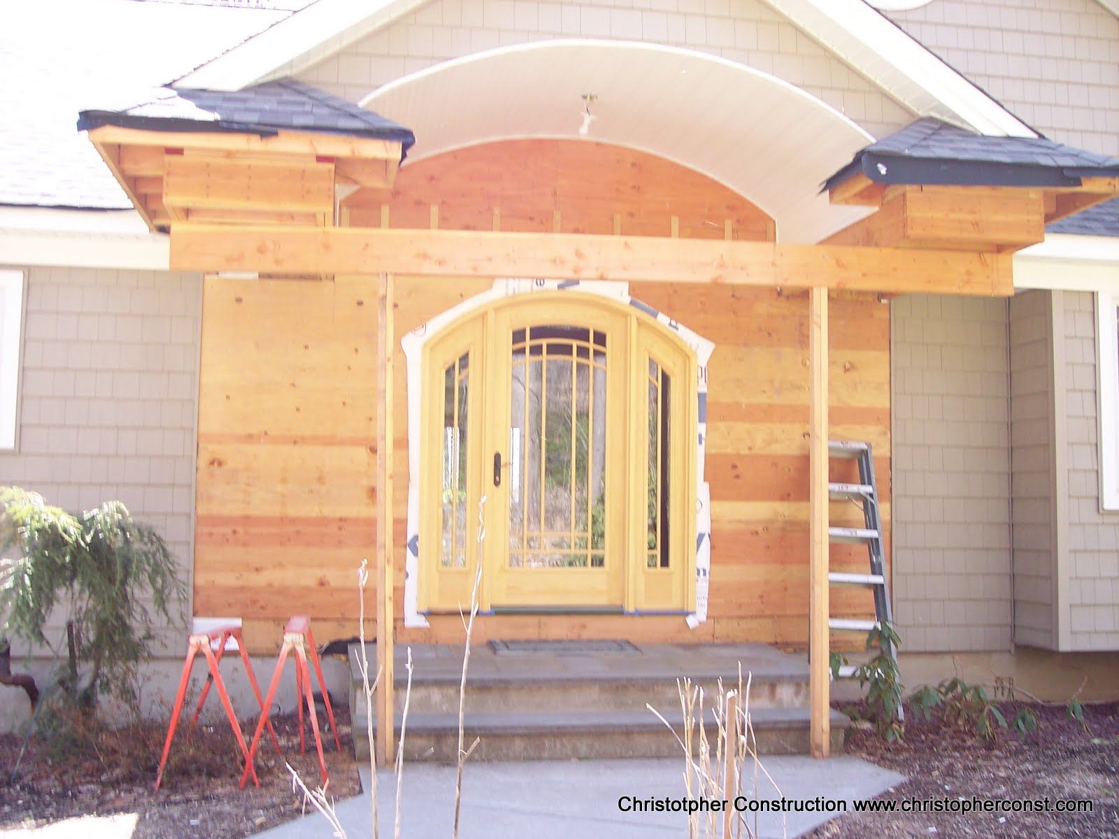 House entrance under construction with plywood framing, yellow double doors, and red sawhorses in front