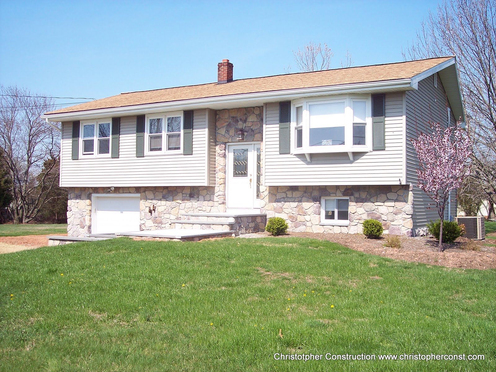 Suburban split-level house with white siding, tan brick, and a green lawn under a blue sky