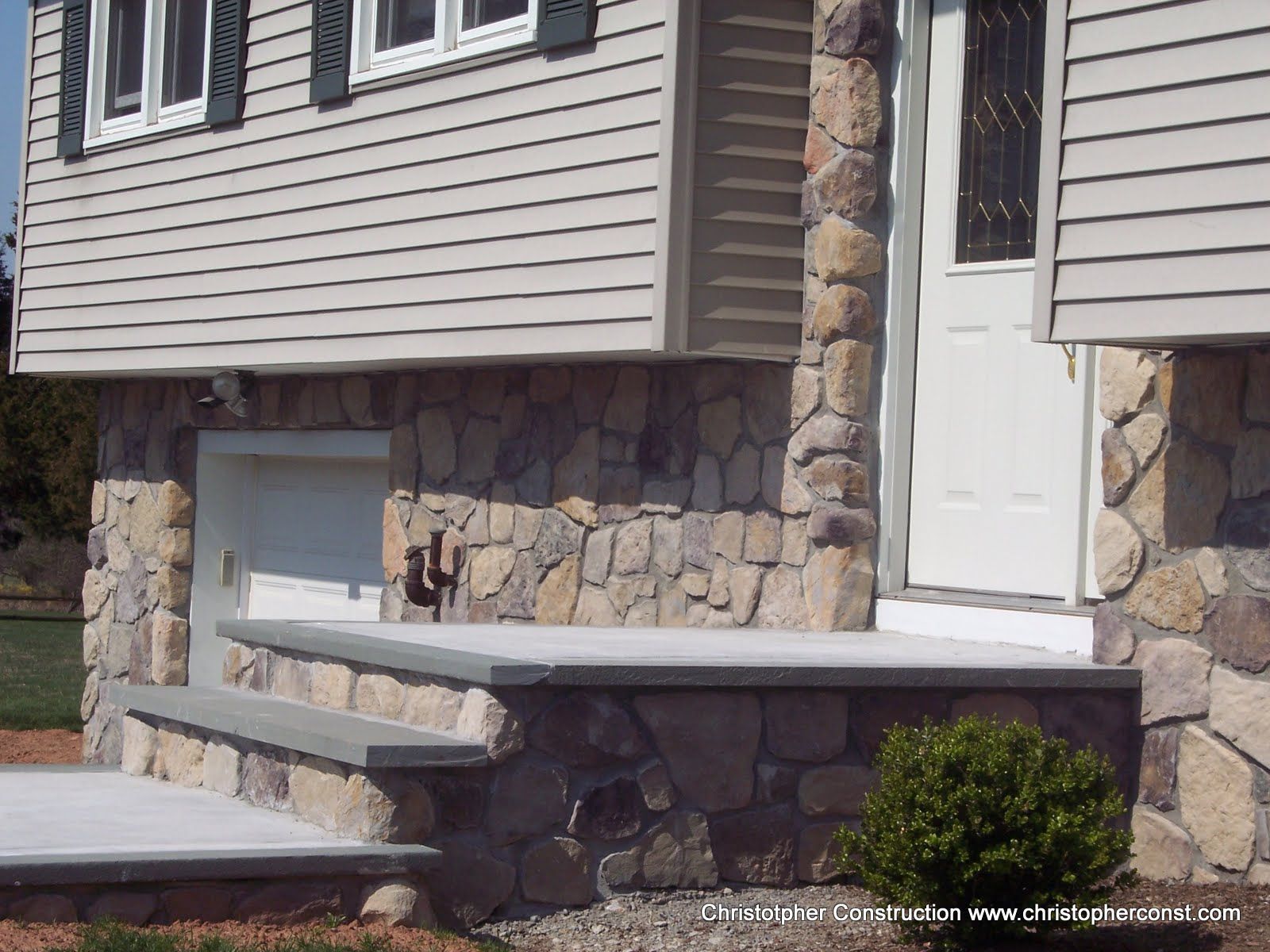 Front porch of a house with stone columns, white door, and light siding, with a concrete step and railing.