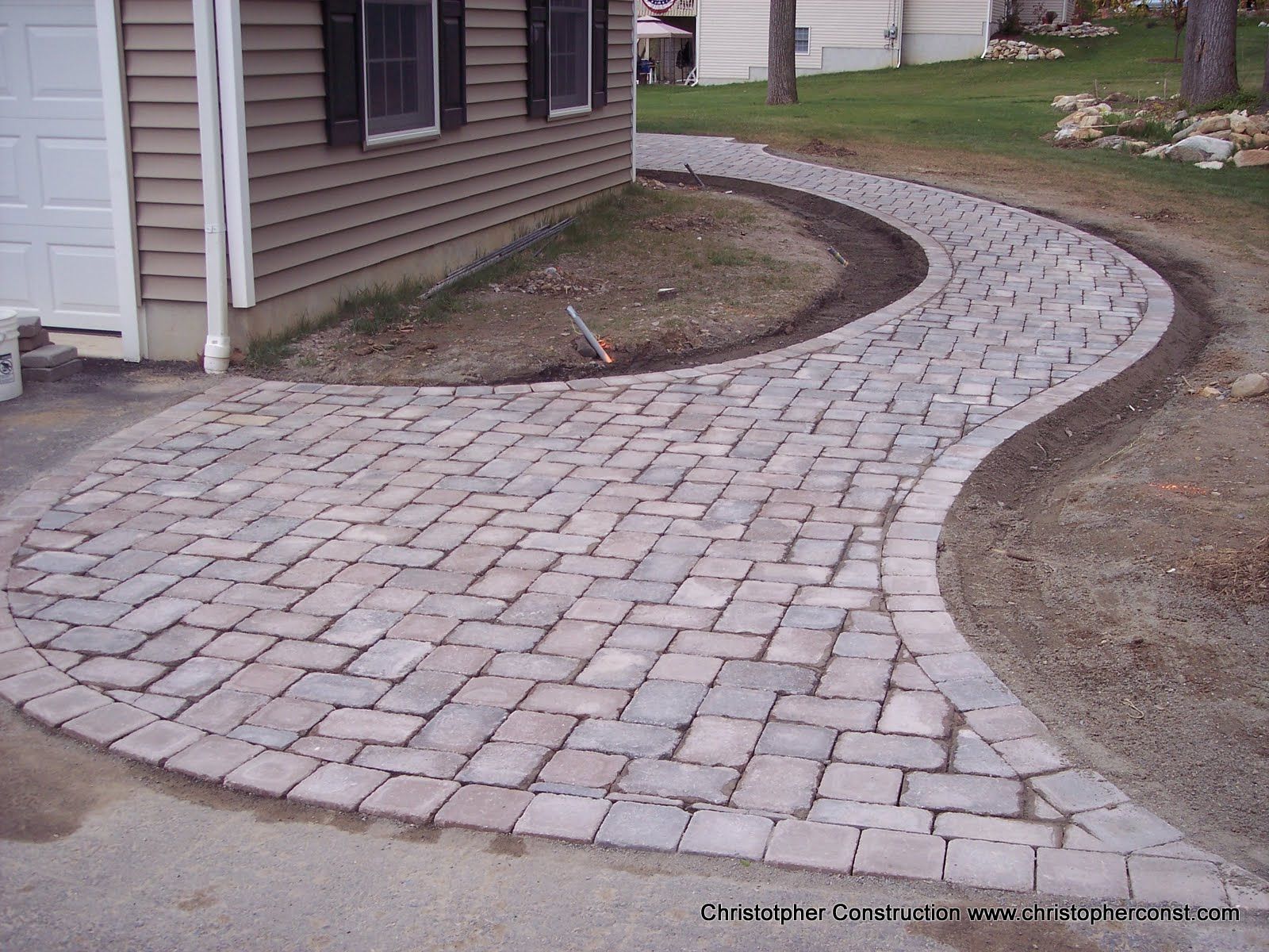 Curved cobblestone walkway beside a house, bordered by dirt and grass.