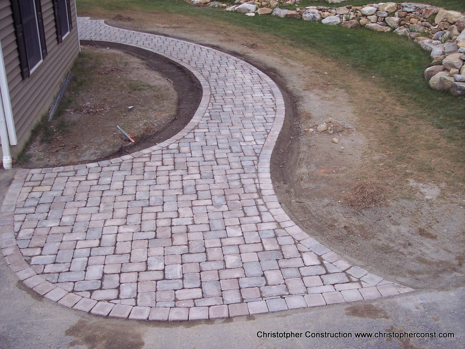 Curving brick walkway beside a house, transitioning to dirt and grass with a stone border.