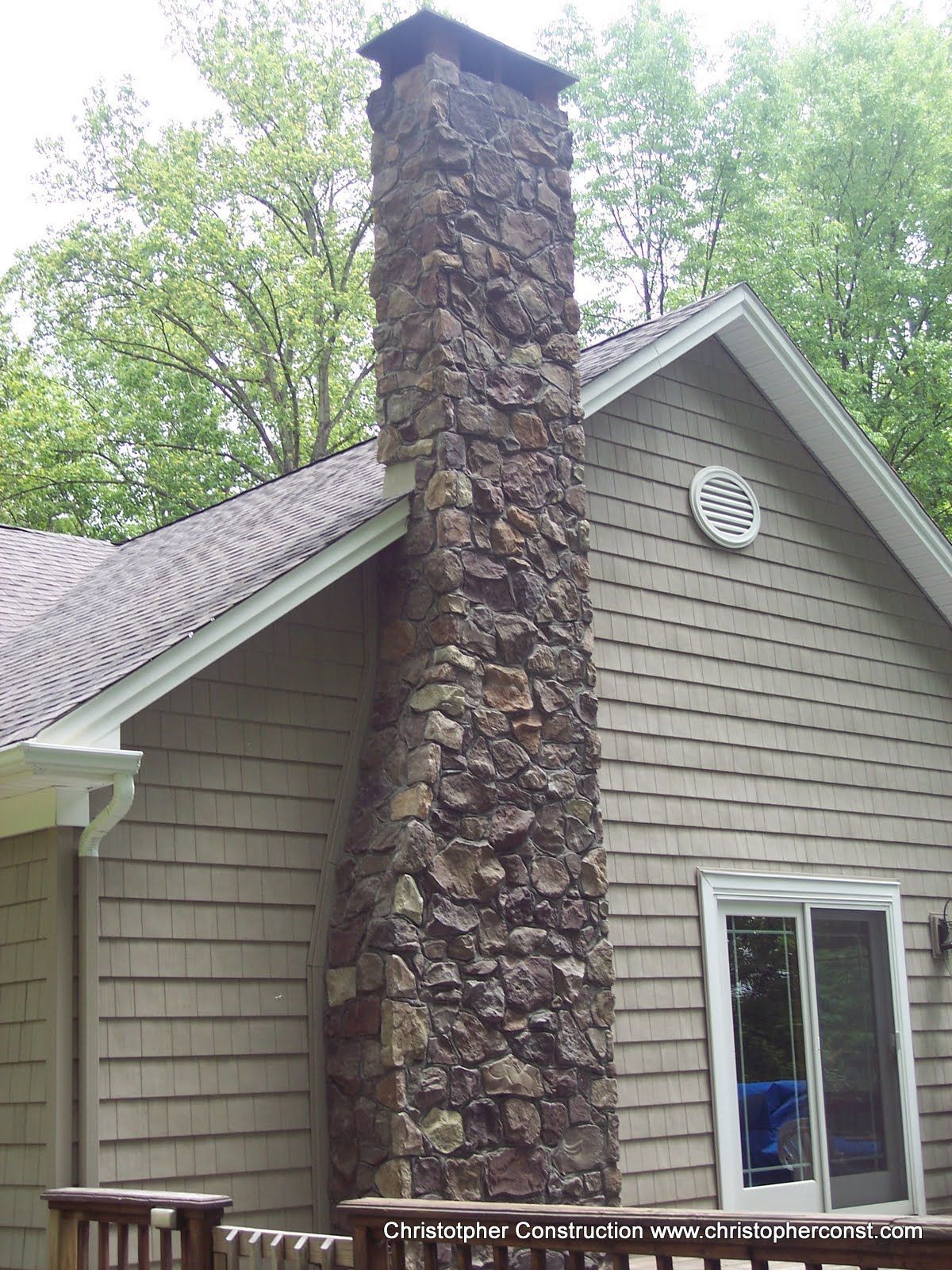 Stone chimney on a house with gray siding and trees in the background