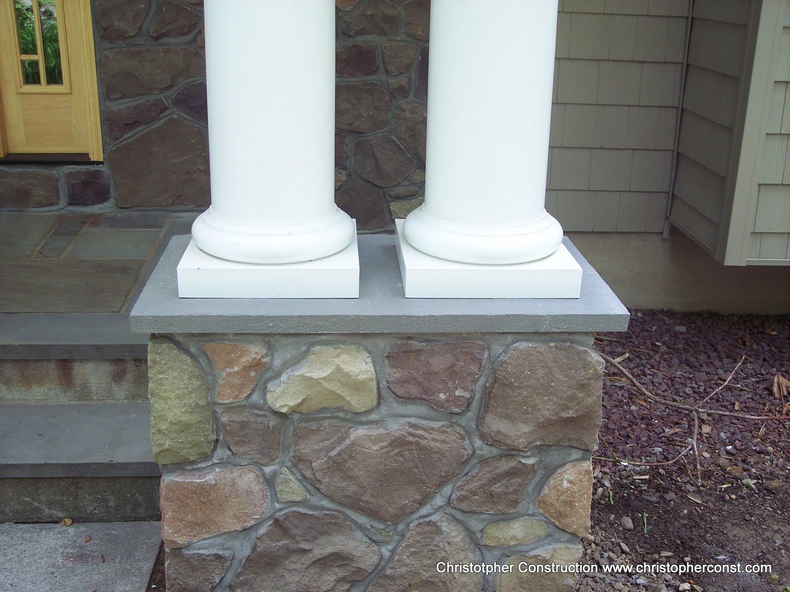 Stone porch with two white columns on a stone base beside steps and siding
