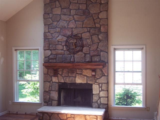 Stone fireplace with mantel in a beige living room, flanked by two windows and a stone hearth.