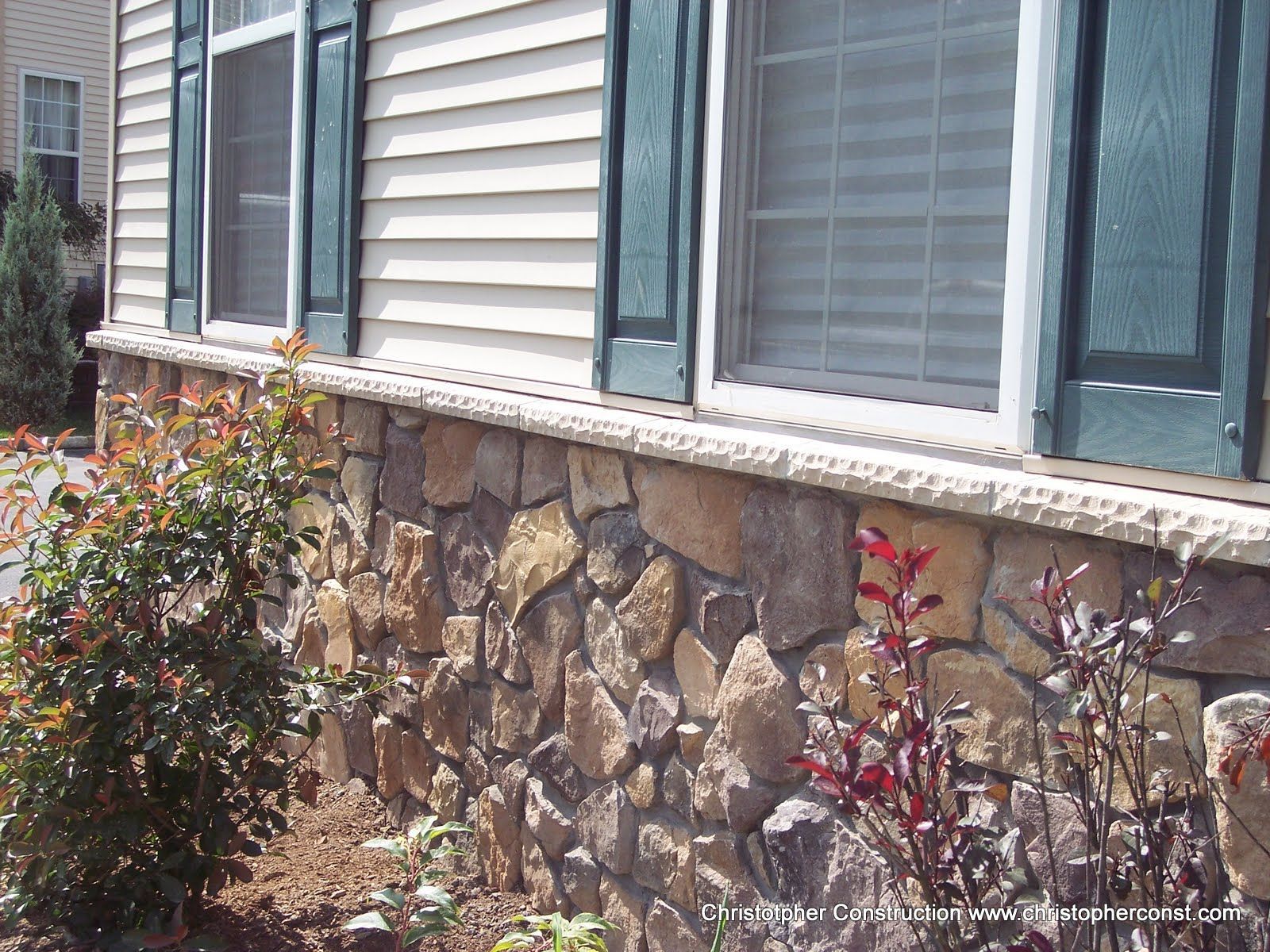 House exterior with stone foundation, white siding, teal shutters, and shrubs along the wall.
