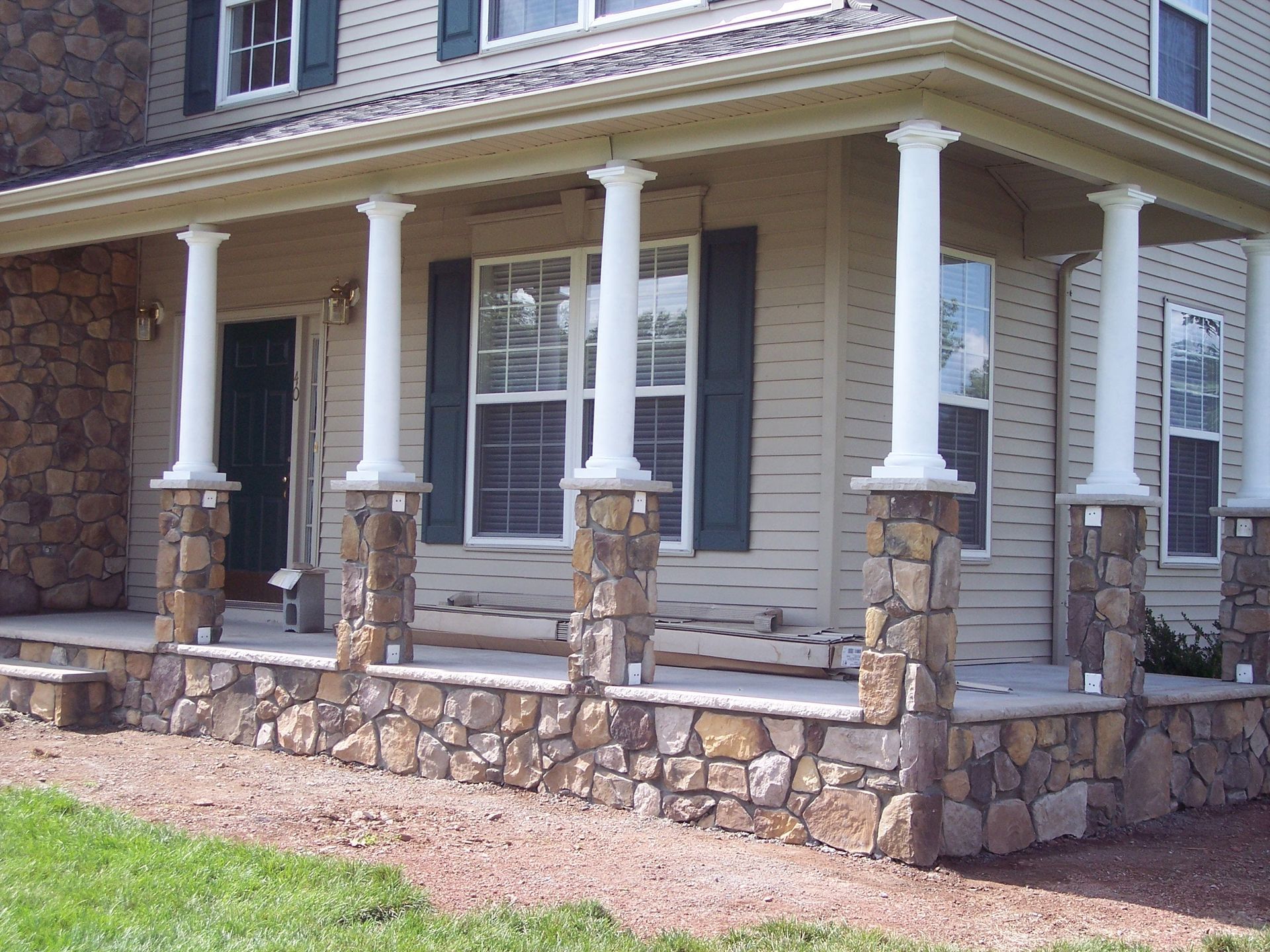 Stone-front porch with white columns on a beige house and brick steps, viewed from the front yard