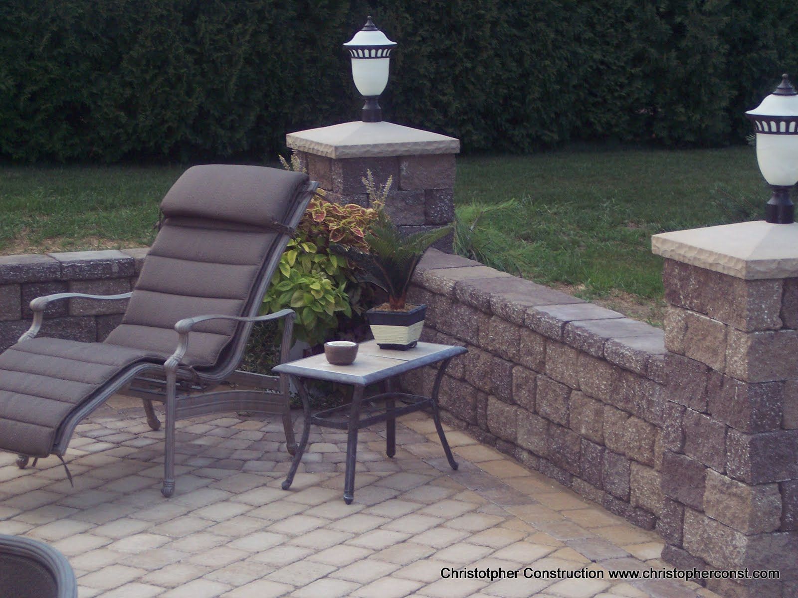 Patio lounge chair beside a small table with potted plants and stone wall lanterns in a backyard.