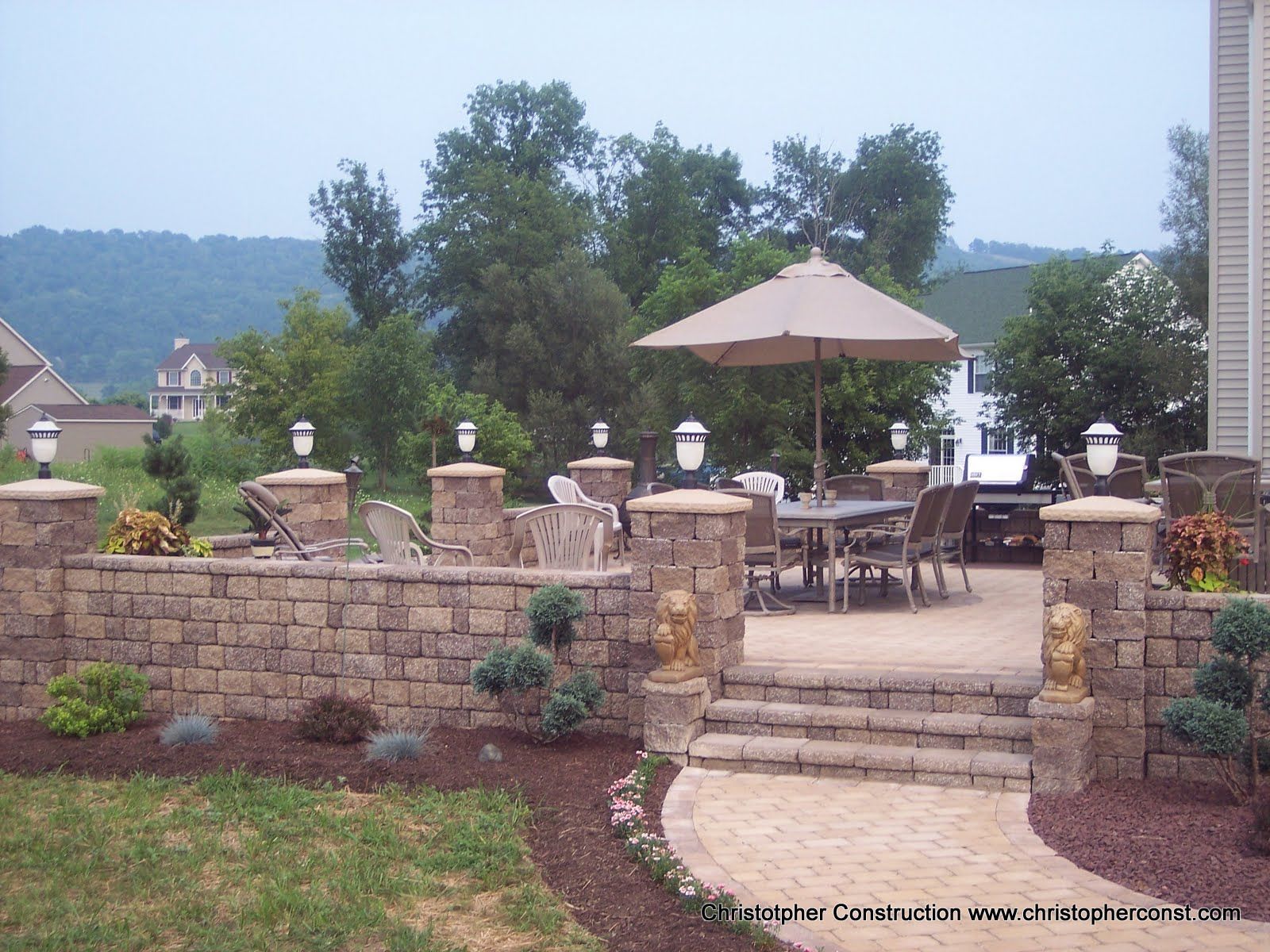Patio with stone wall, tables, umbrellas, and chairs overlooking a wooded valley under a clear sky