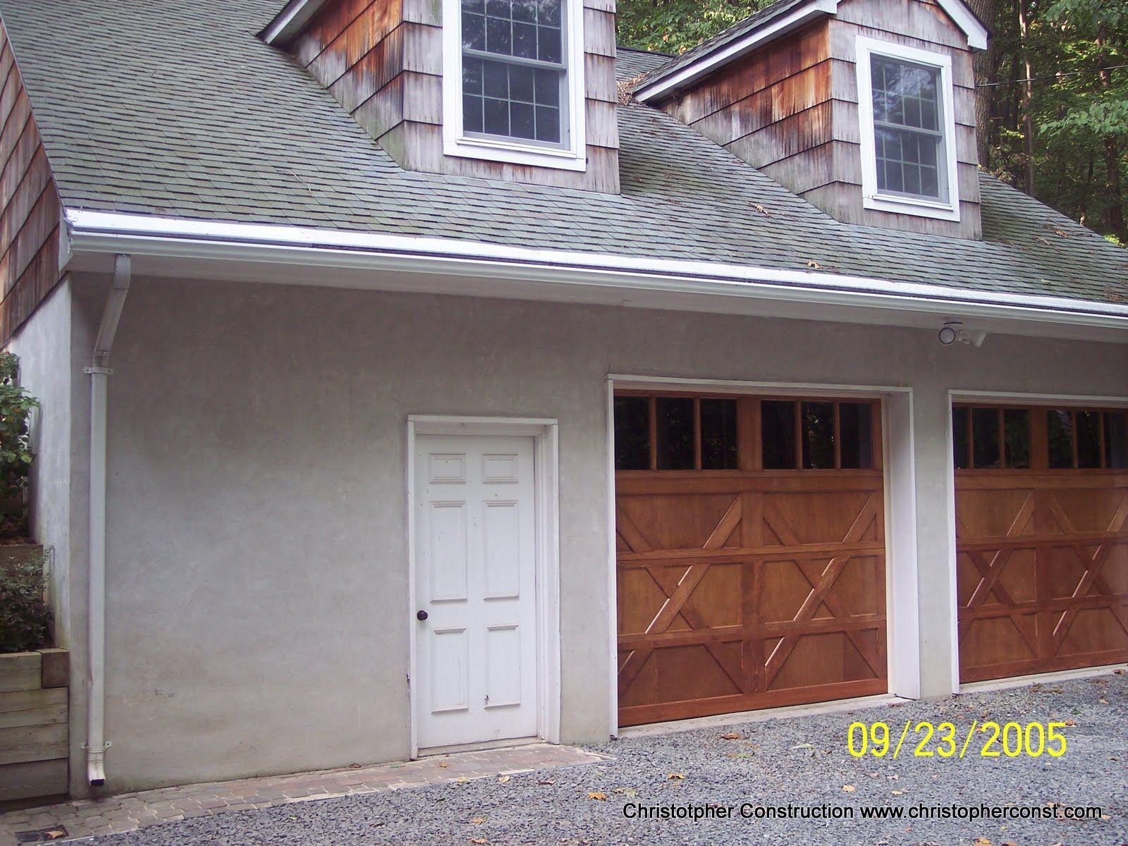 Gray house exterior with a white side door and double wooden garage doors under a shingled roof.