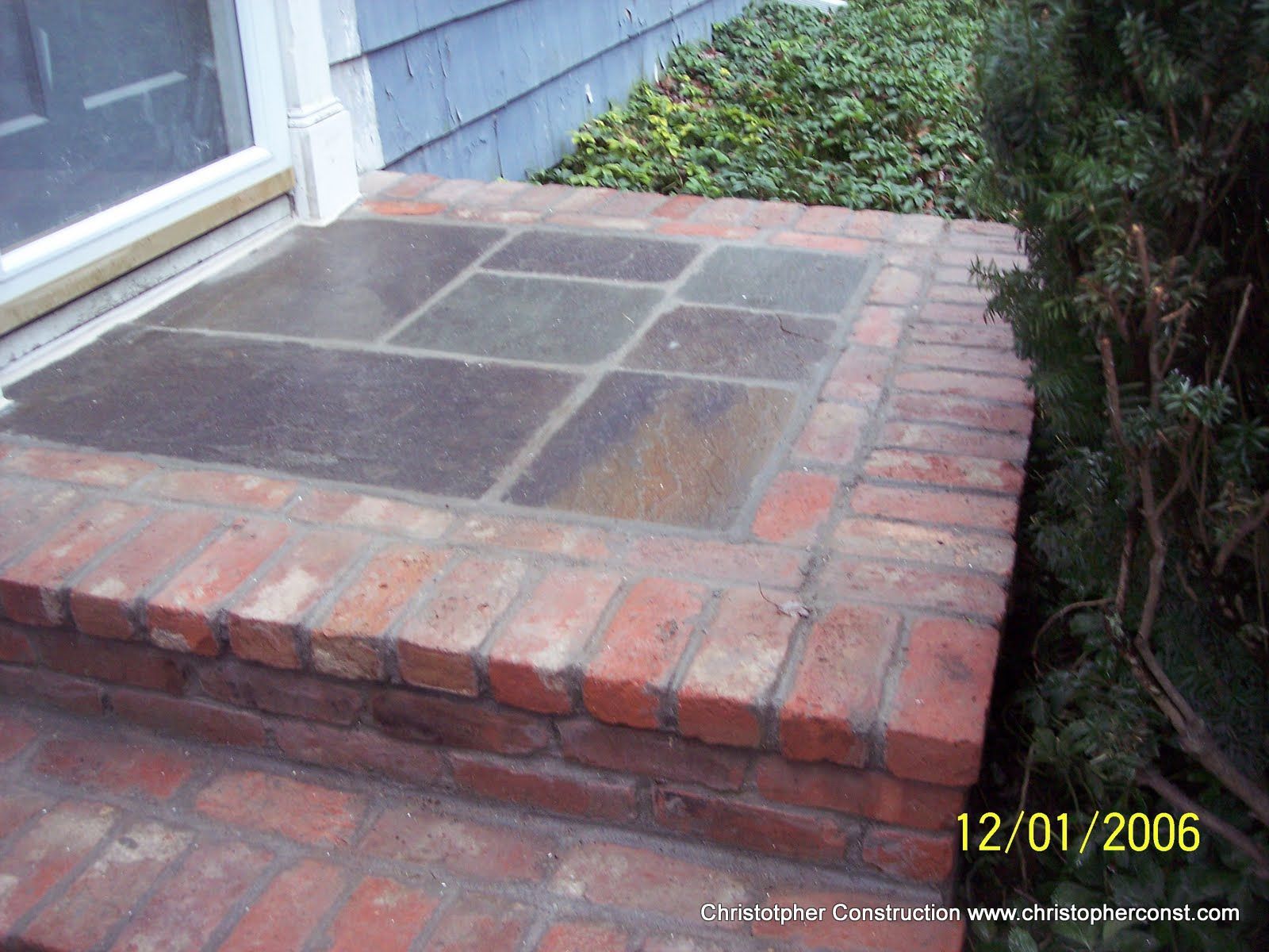 Brick front step with dark square pavers beside a house and shrubbery
