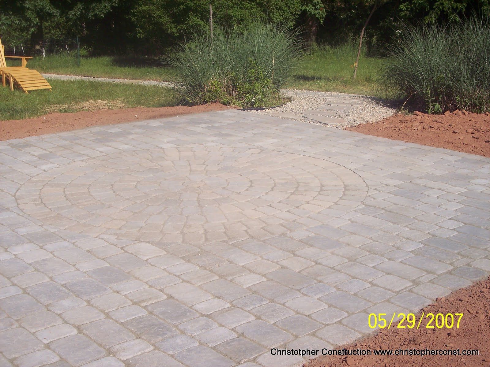 Empty stone-paver patio in a landscaped yard with shrubs and grass.