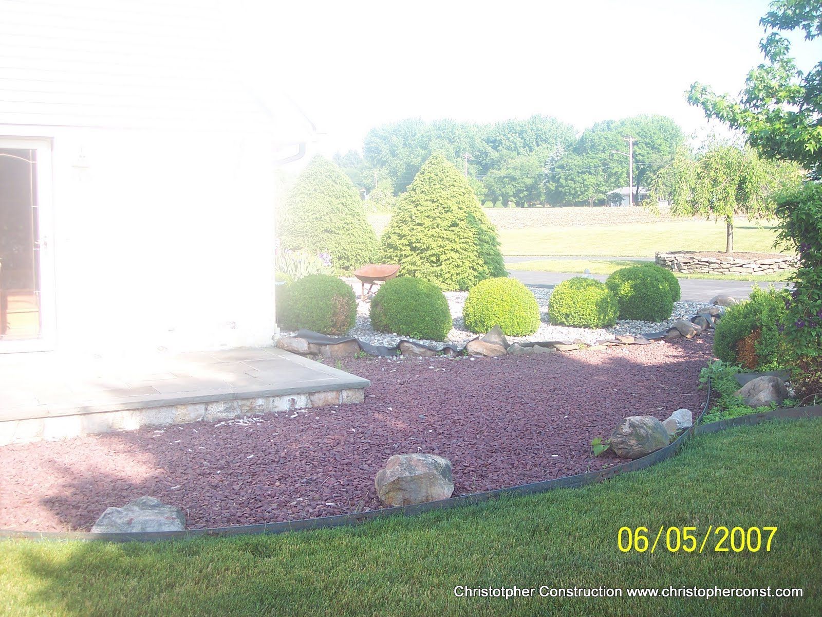 Sunny landscaped yard with shrubs, rocks, and red mulch beside a white house