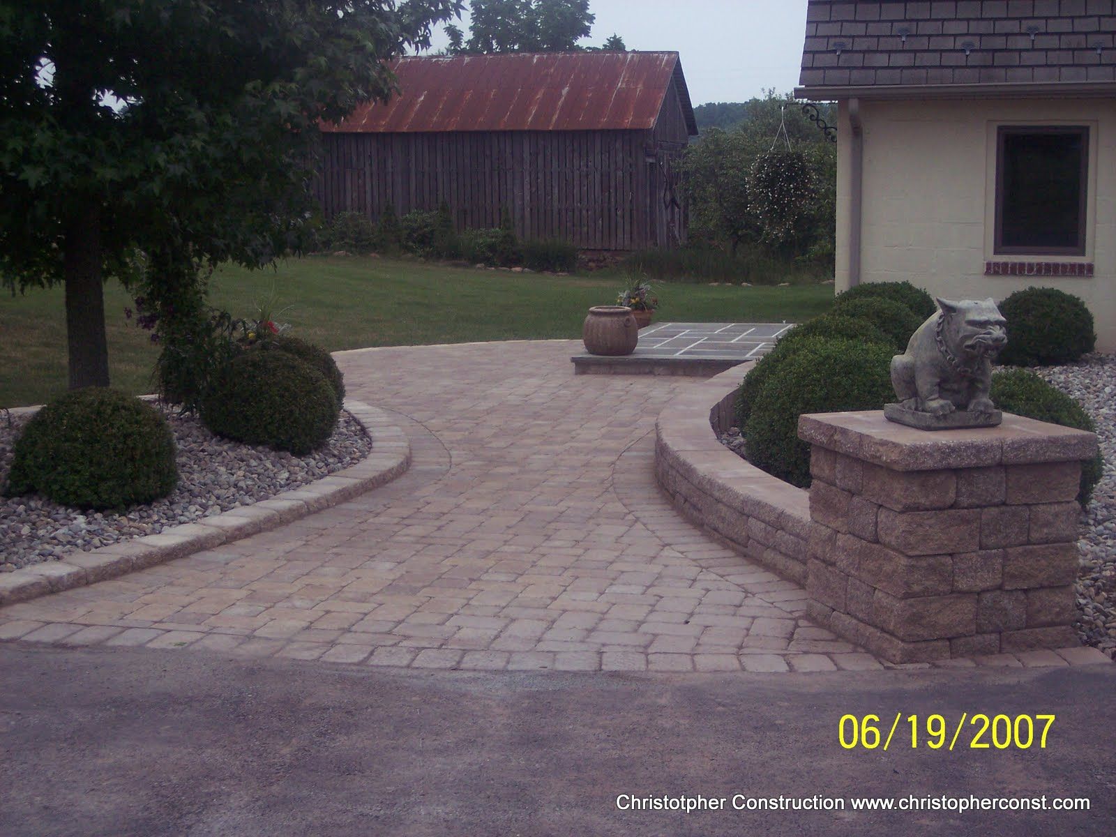 Stone-paved courtyard with curving paths, shrubs, a planter, and a house and barn in the background.