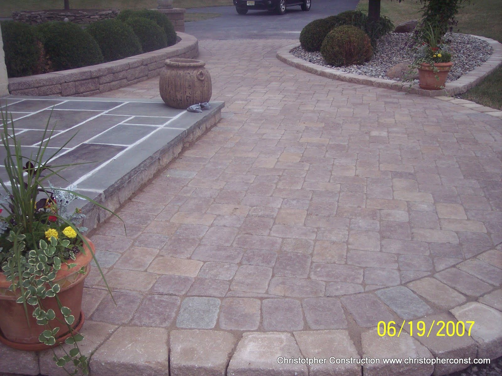 Brick driveway and stone patio with potted flowers, shrubs, and a large urn planter outdoors.