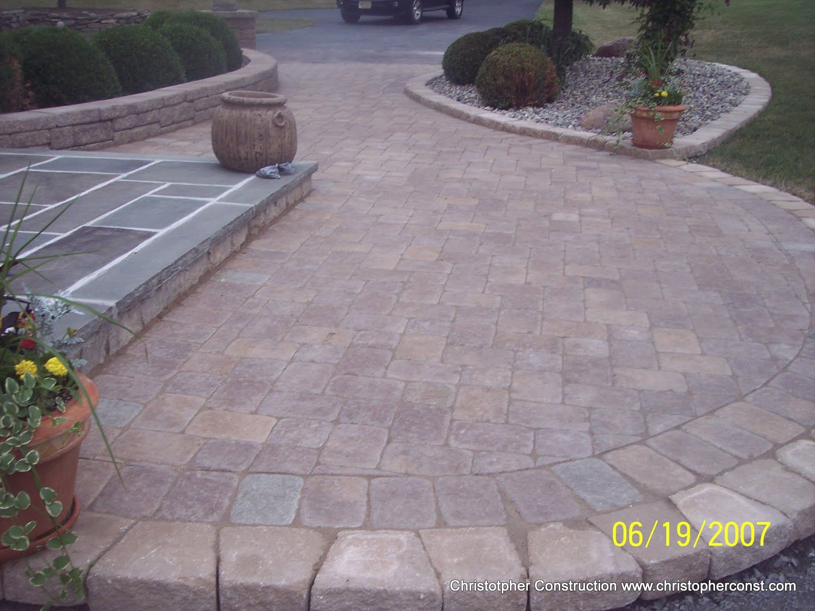 Paved driveway with curved brick border, potted plants, and a small decorative barrel planter.