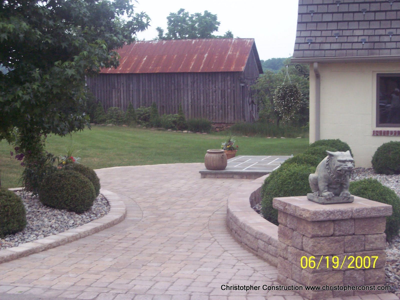 Curved stone walkway beside a house, leading to a lawn and red barn in the background.