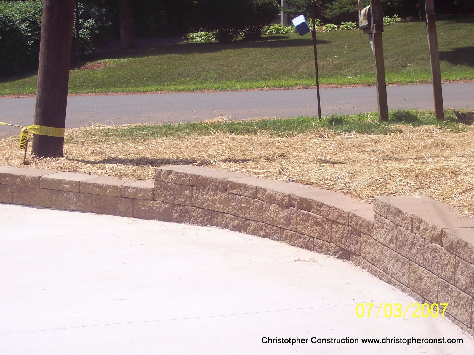 Curved concrete retaining wall beside a sidewalk, with gravel, grass, and a road in the background