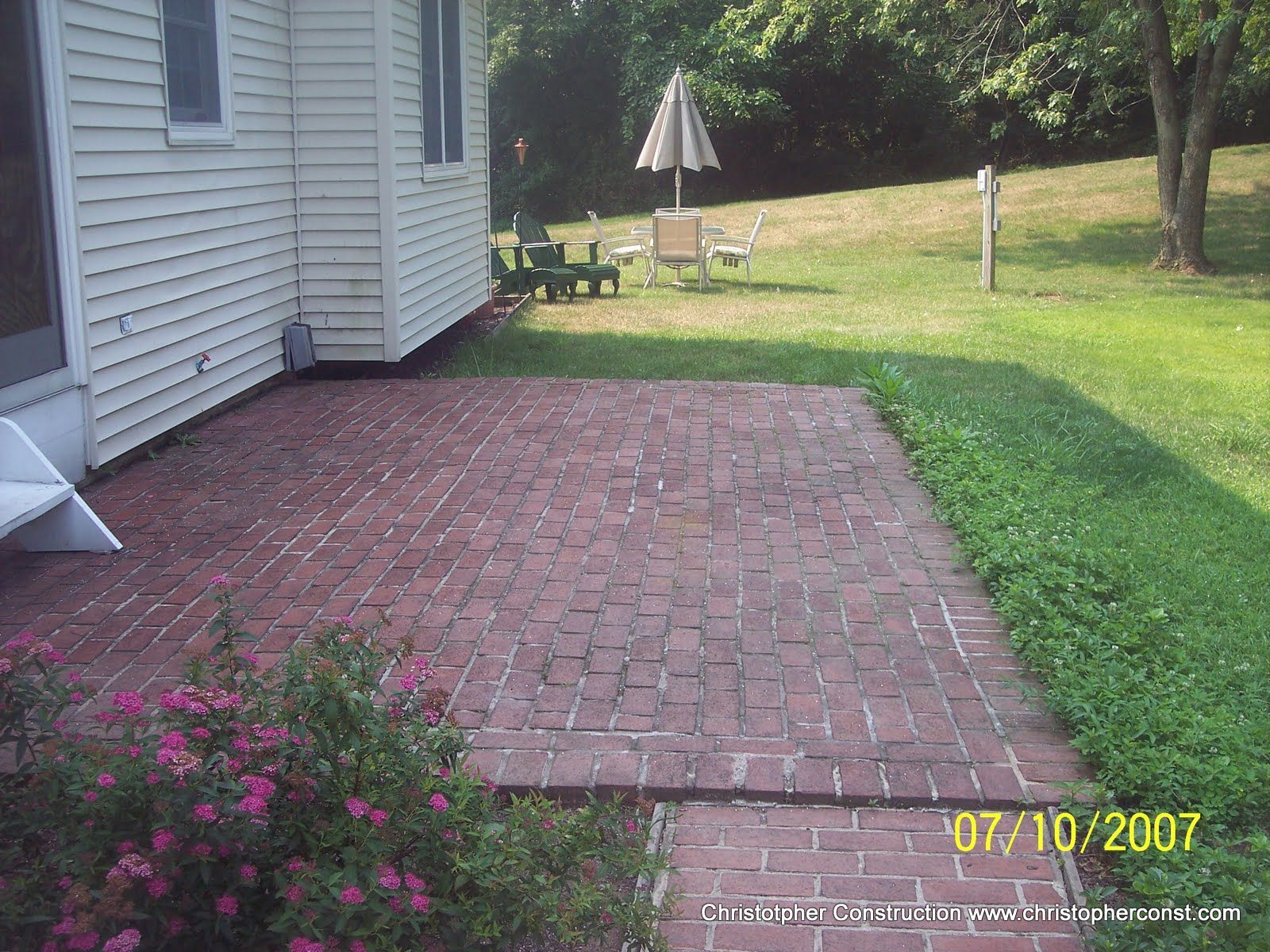 Brick patio beside a white house, opening to a grassy yard with a patio table and umbrella.