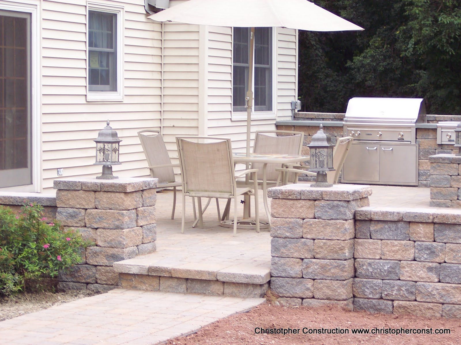 Patio with stone walls, outdoor table and chairs, grill, and large umbrella beside a house