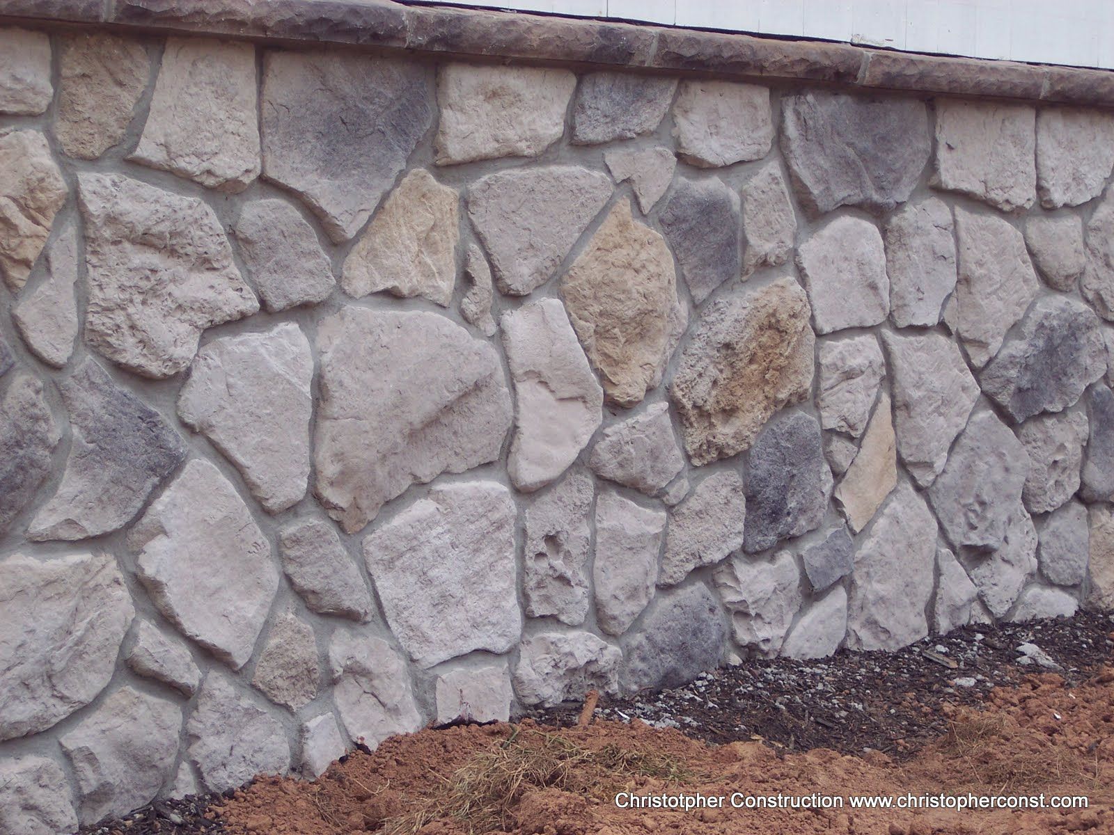 Stone wall with irregular gray and beige rocks and a sloped concrete top edge.