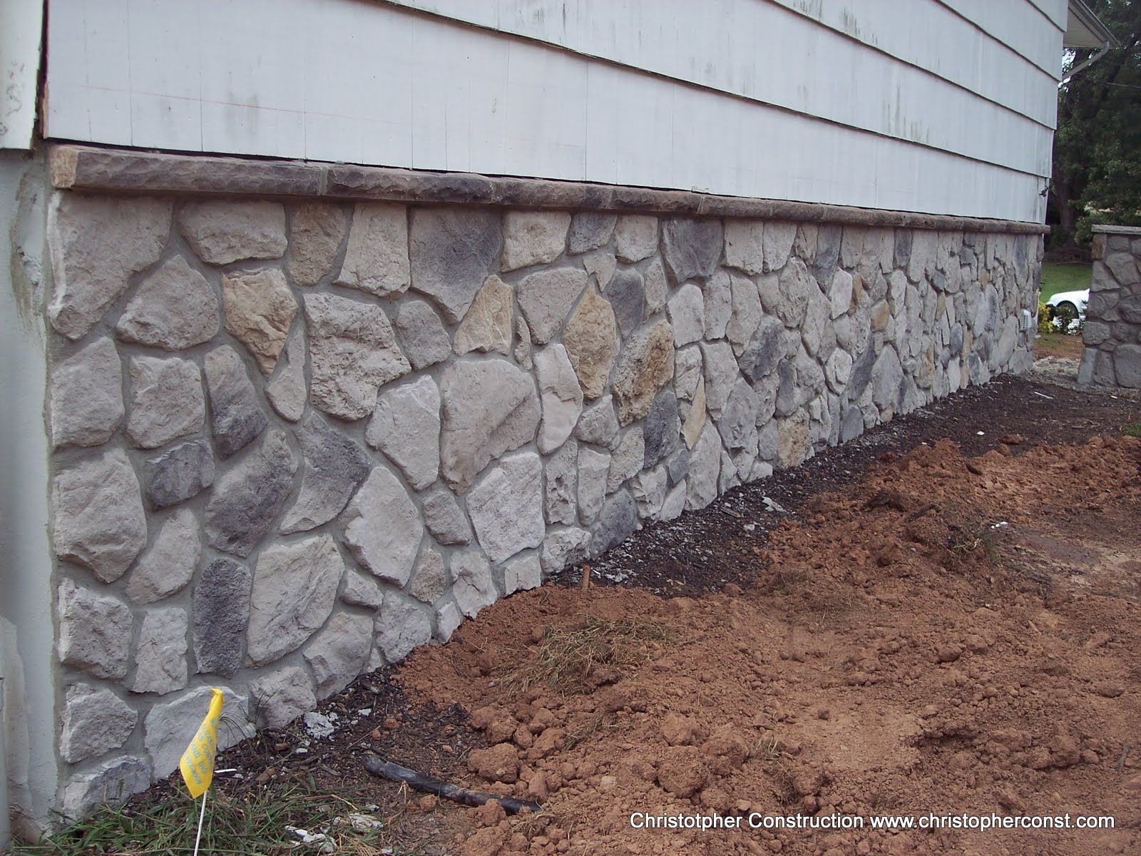 Stone foundation wall beside a house with fresh soil along the base and a yellow marker flag