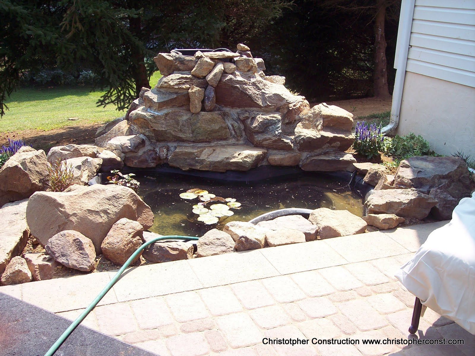 Backyard rock fountain with a small pond, bordered by stones beside a patio and house wall