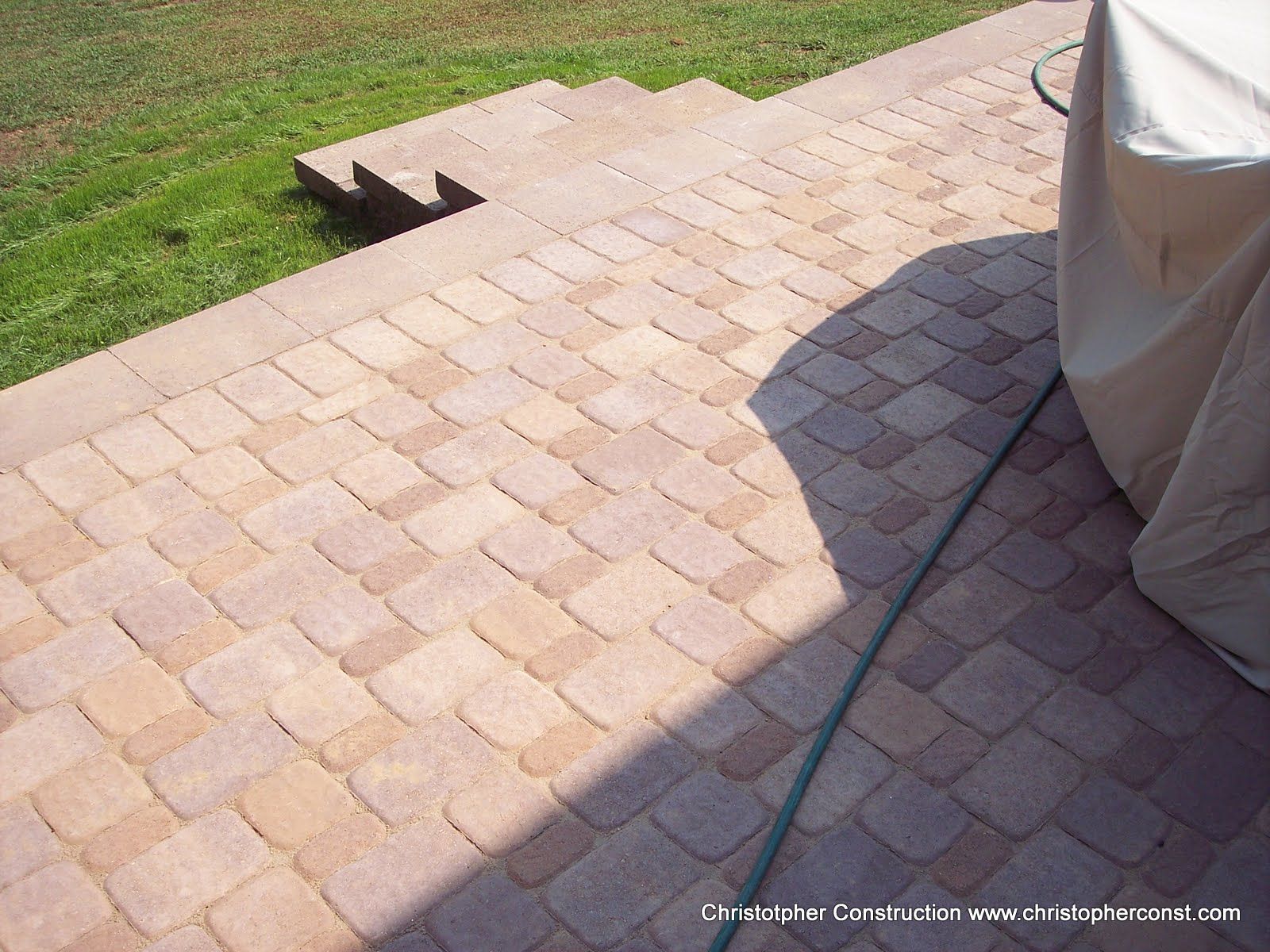 Brick paver patio beside a grassy lawn, with a step and a covered object casting a shadow.