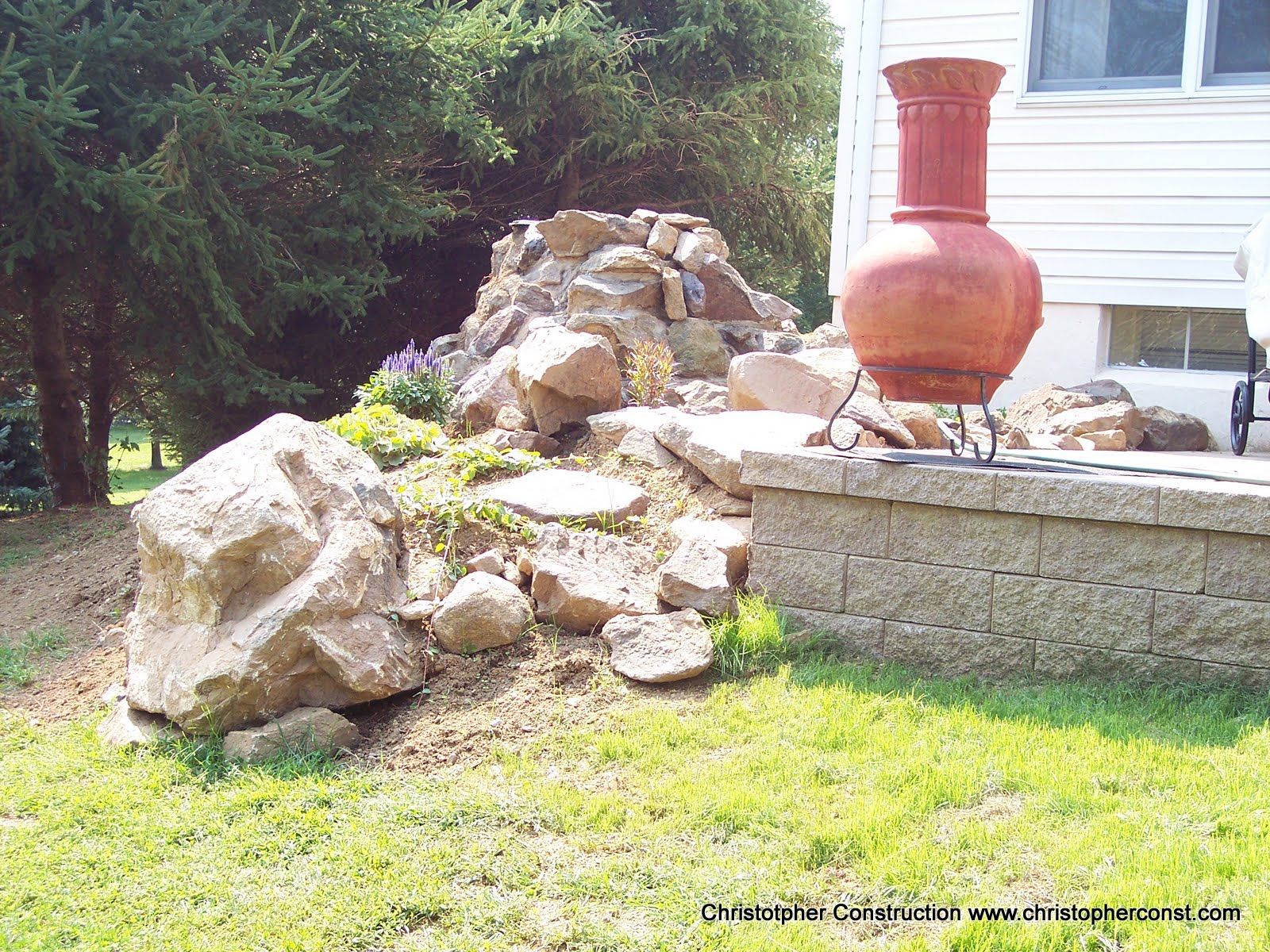 Large red clay urn on a stone ledge beside a rock garden and house exterior