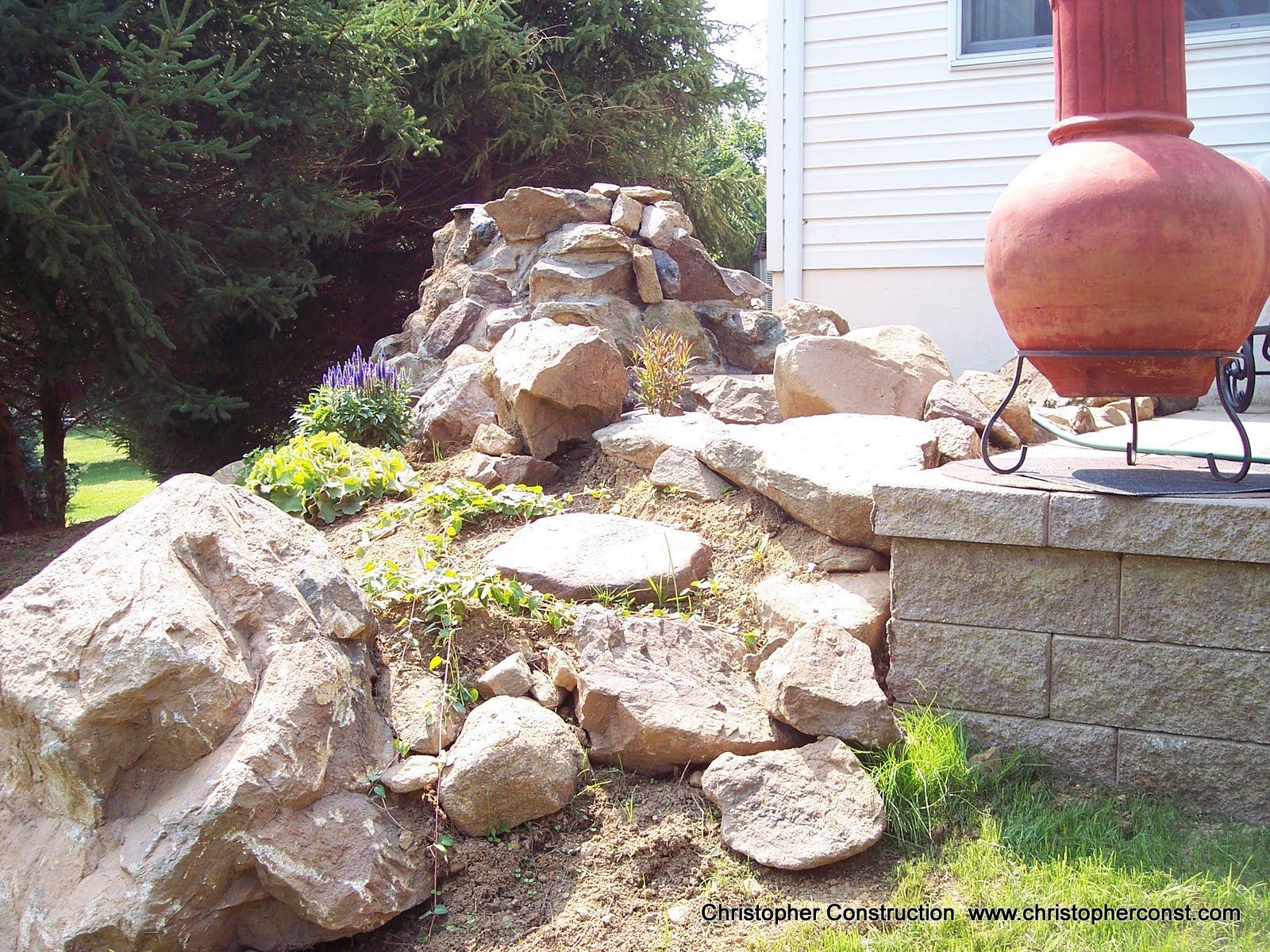 Rock garden with large boulders beside a house and a red chimney pot outdoors.