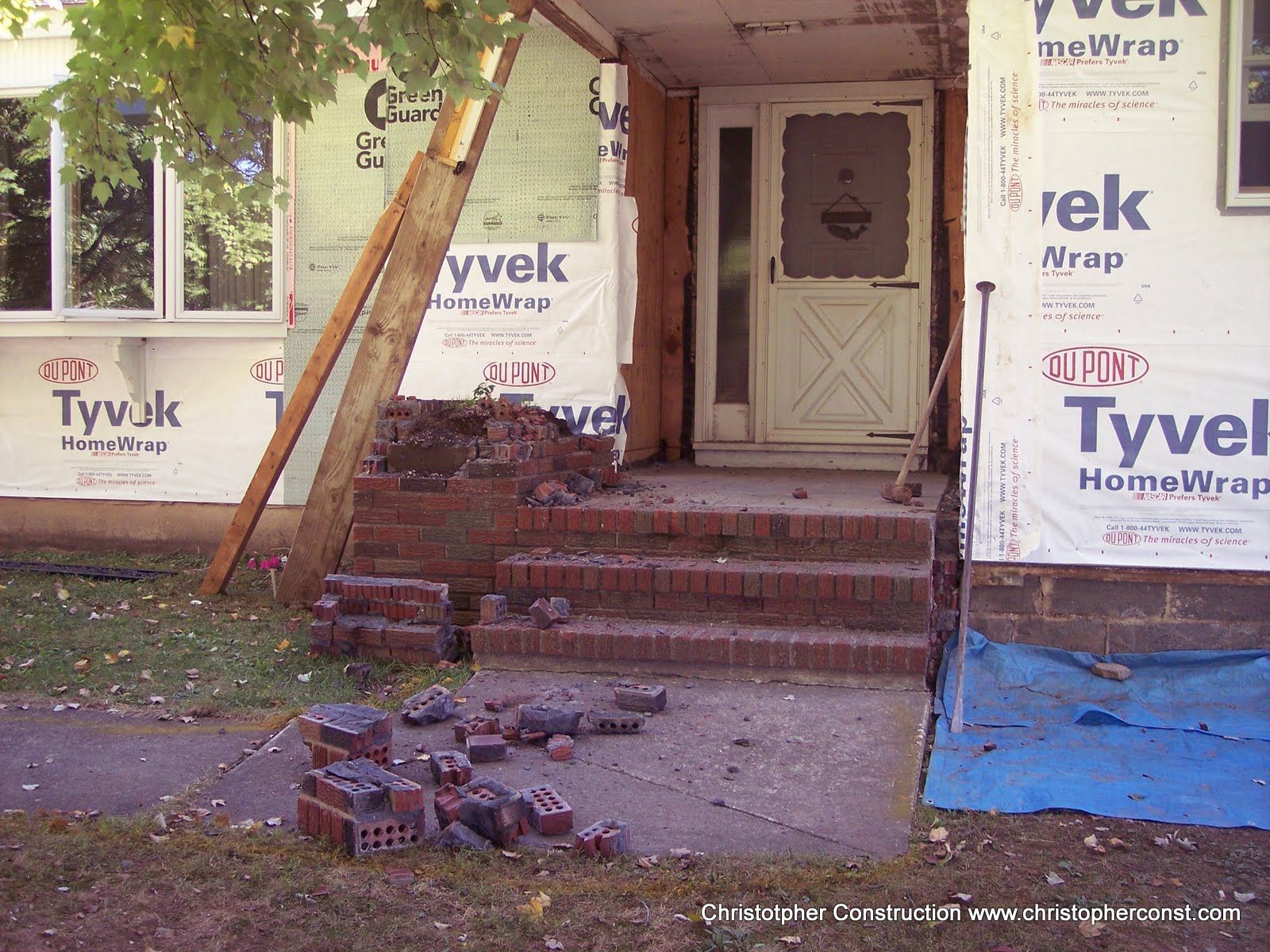 Front porch of a house under renovation with Tyvek wrap, steps, and a closed front door