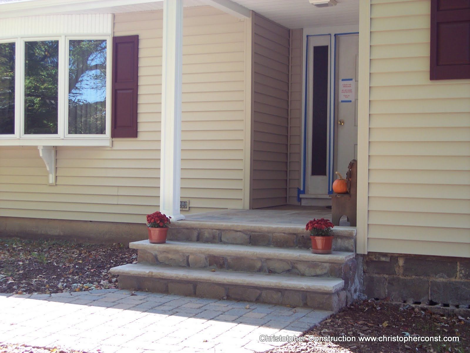Front porch with beige siding, stone steps, white columns, and two red flower pots by the door.