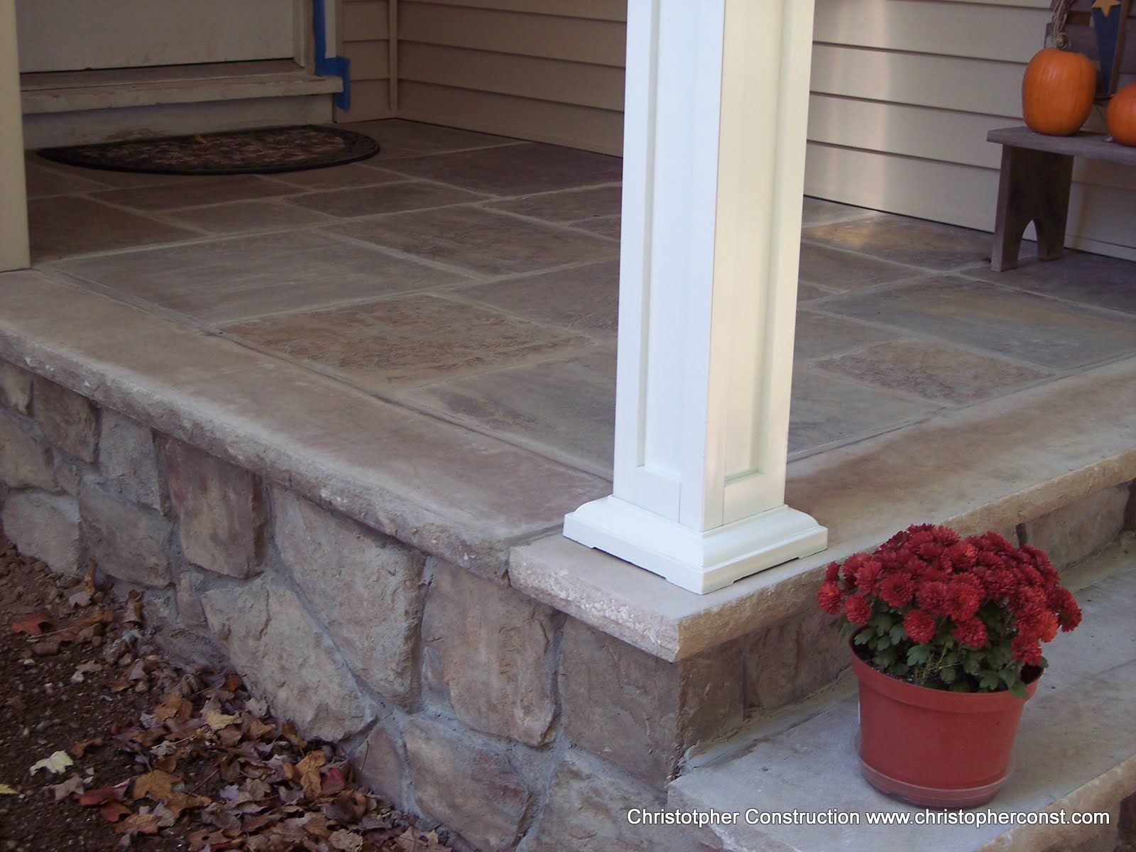 Stone porch with white column, autumn leaves, and a red potted mum by the steps
