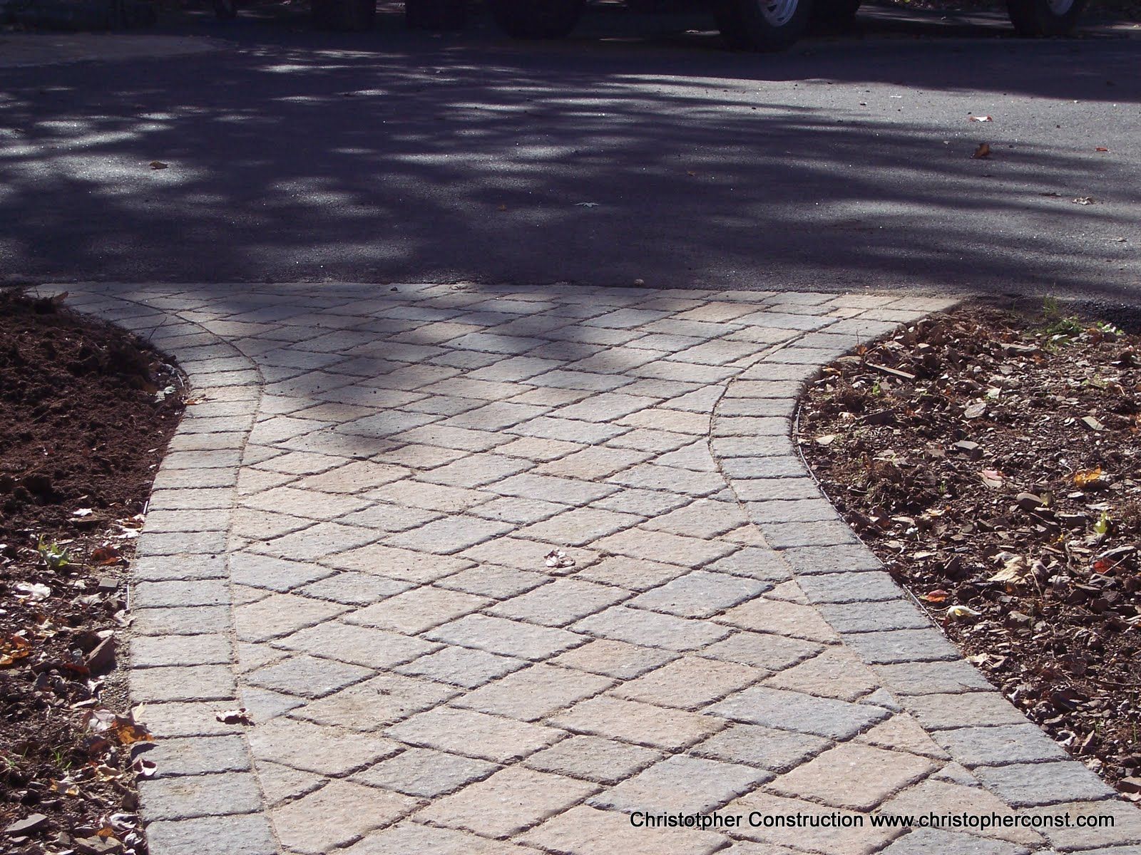 Curved brick walkway leading into a shaded driveway with mulch on both sides
