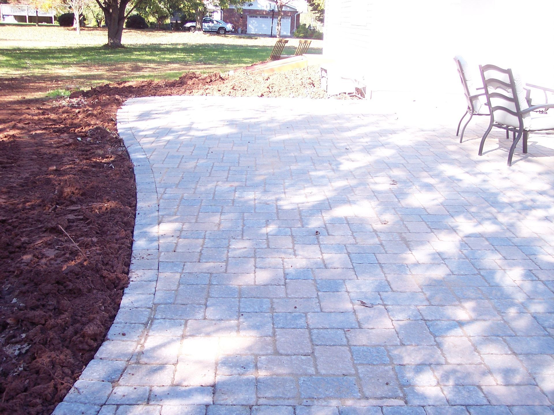 Curved stone patio beside a grassy yard, with a dark mulch border and chairs in the sunlight.