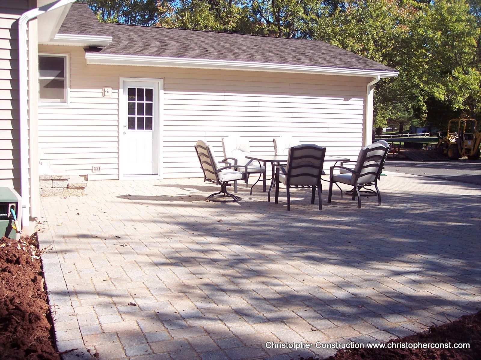 Patio with several chairs and a glass table beside a white house on a paved yard
