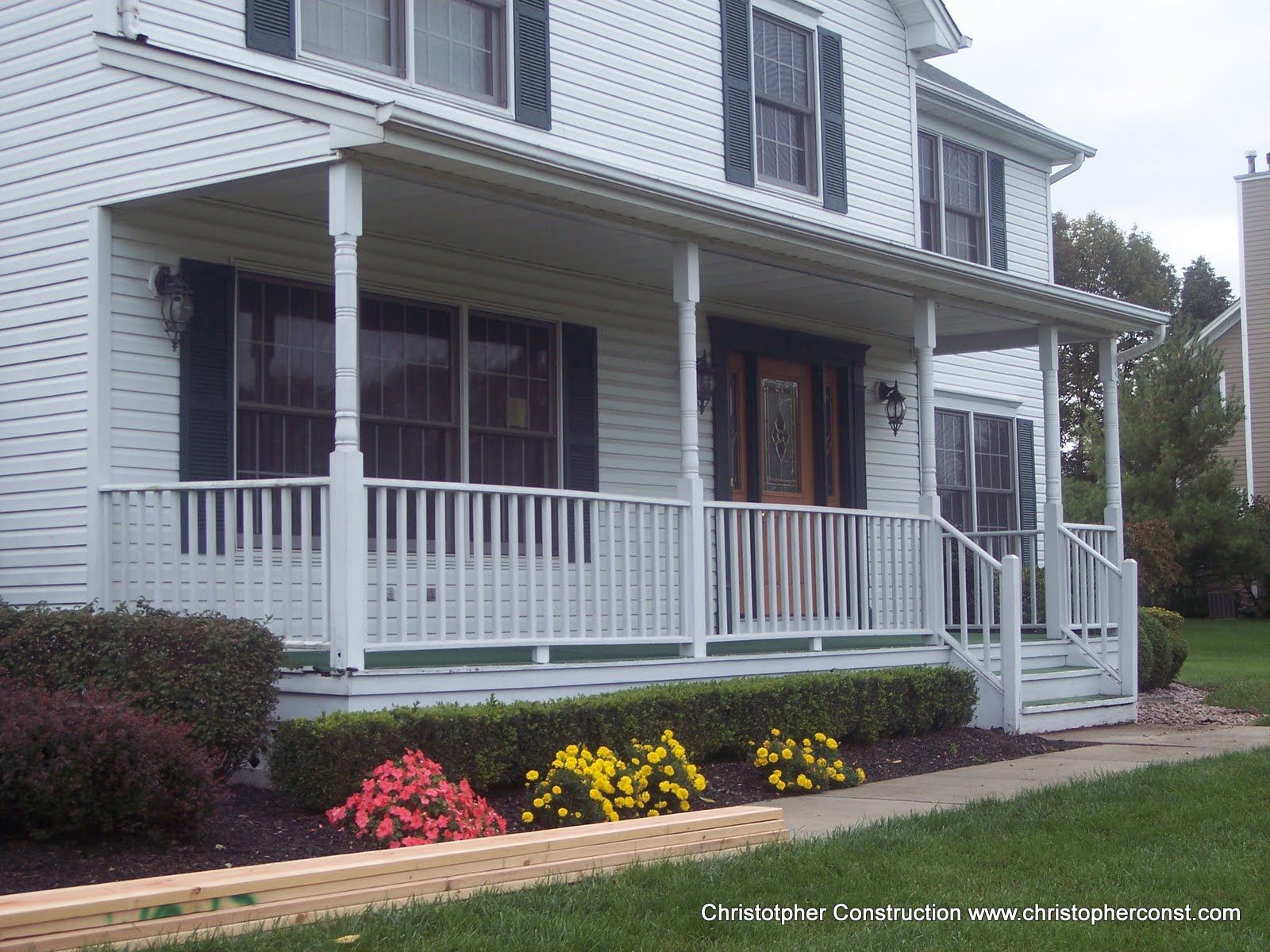 White house with a covered front porch, railings, steps, and pink flowers in the yard