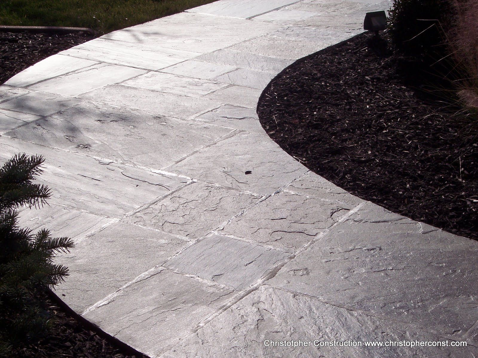 Curved concrete sidewalk bordered by dark mulch and bushes, with sunlight and shadows across the path