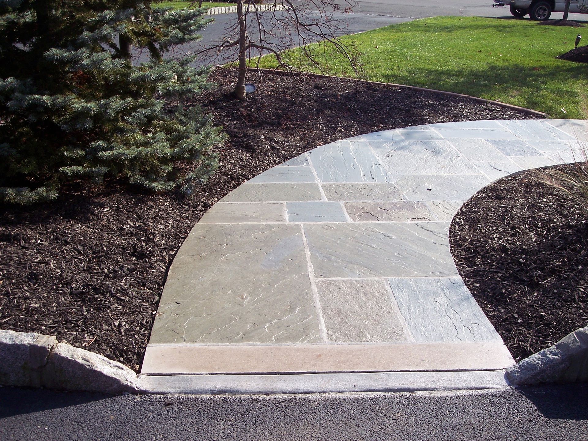 Stone walkway curves through a landscaped garden with mulch and grass.