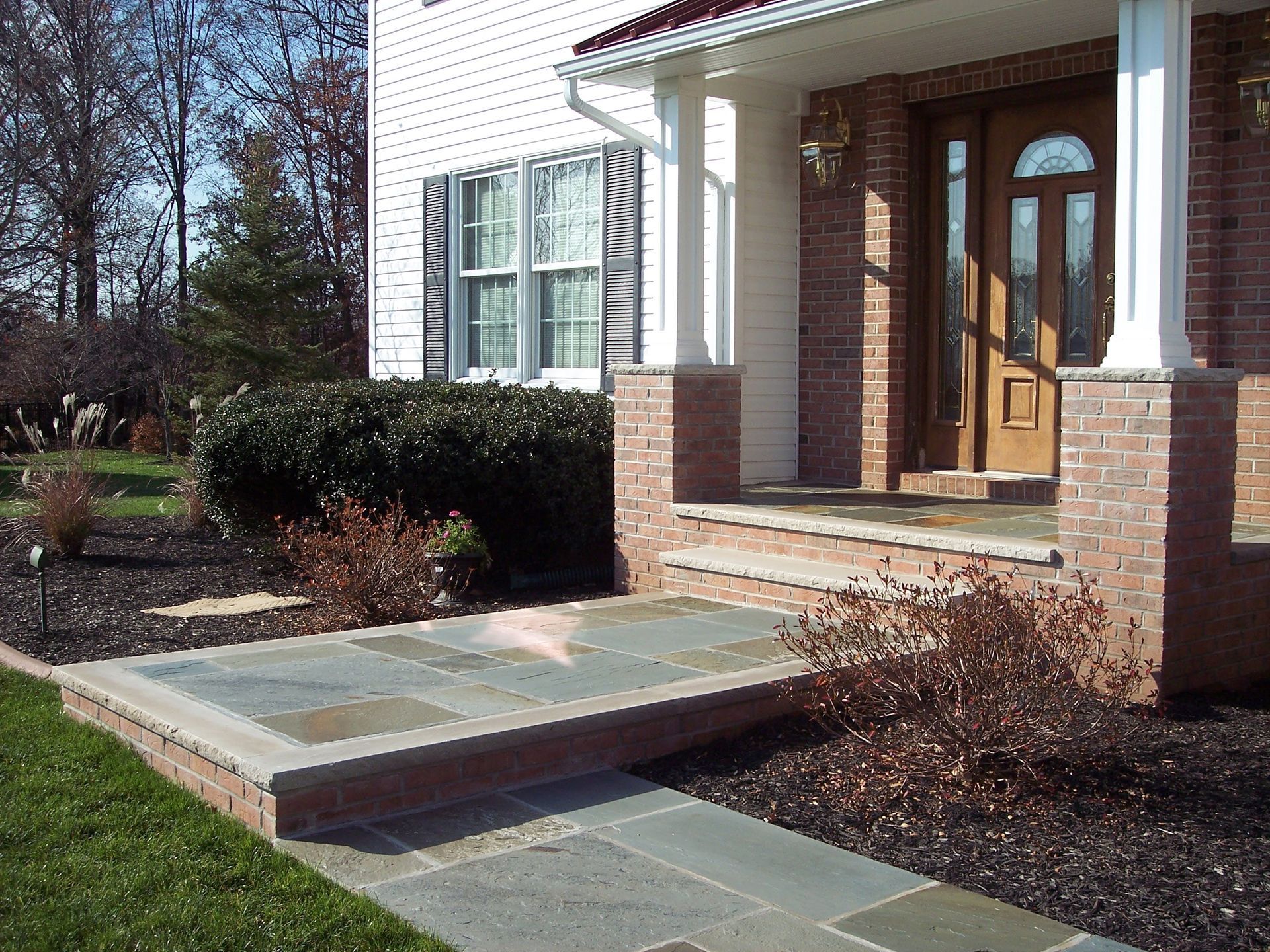 Front porch with a wheelchair ramp leading to a brick house entrance, bordered by shrubs and walkway.