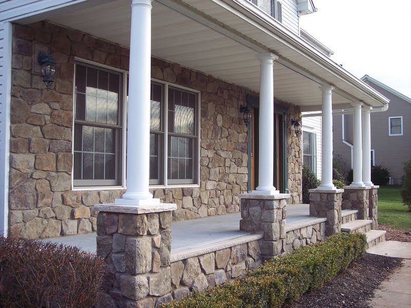 Stone-front house porch with white columns and windows, beside a sidewalk and trimmed shrubs
