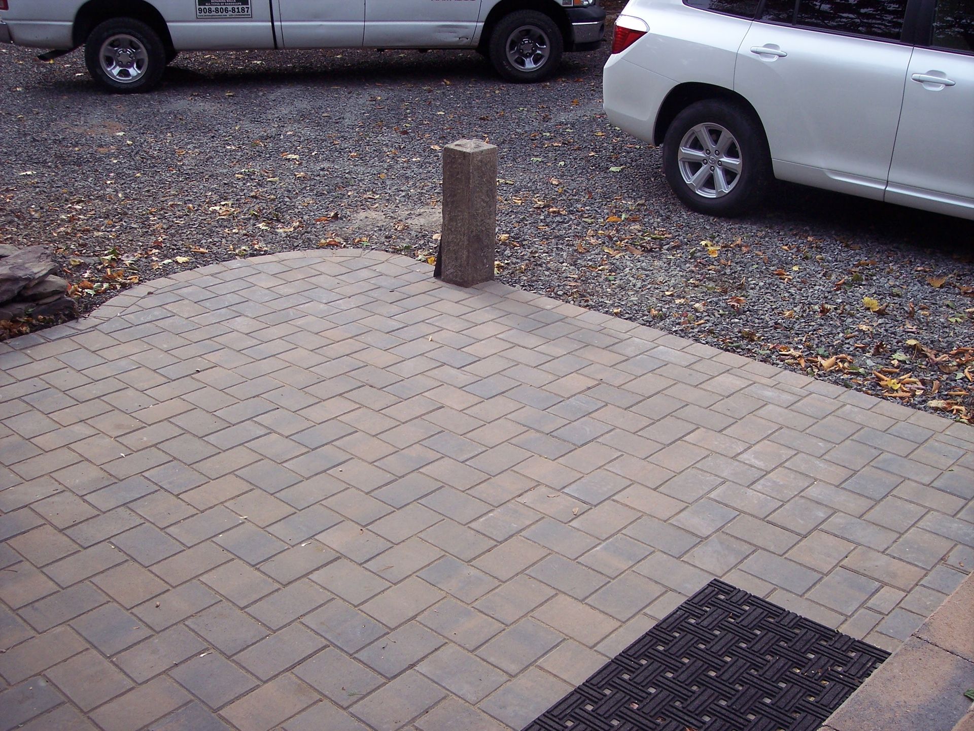 Brick-paved sidewalk with a concrete bollard, gravel parking area, and parked cars in the background
