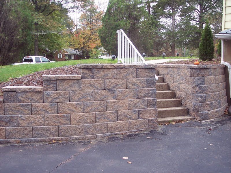 Stone retaining wall with built-in steps leading up to a yard, beside a driveway and trees