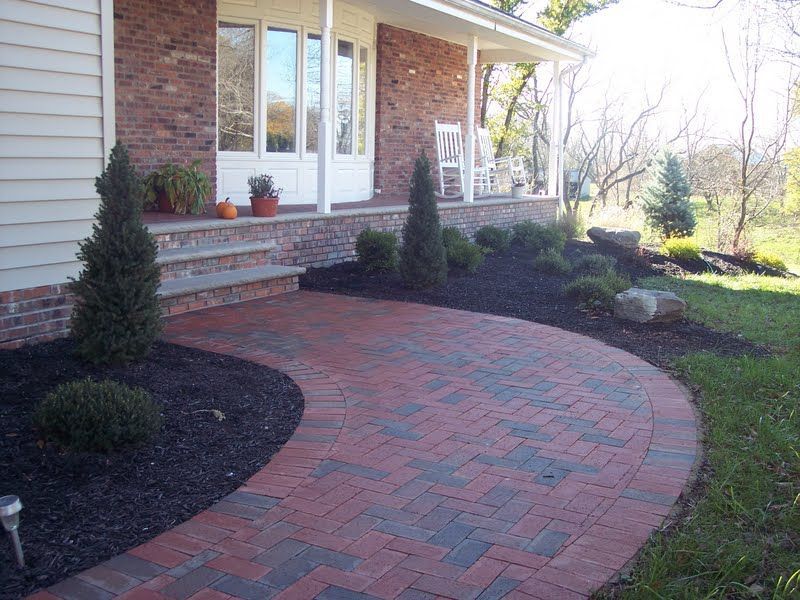 Curved red brick walkway leading to a brick house porch with potted plants and shrubs