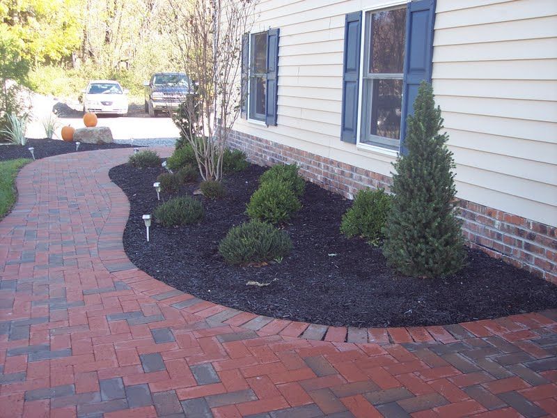 Front yard brick walkway curving past landscaped shrubs and a house with beige siding