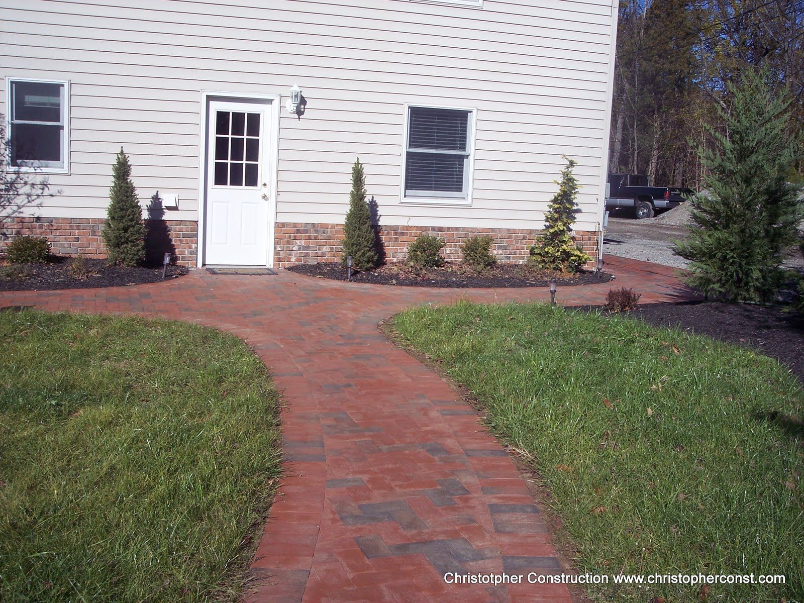 Brick walkway leading to a white door on a house, with green lawn and shrubs on both sides.