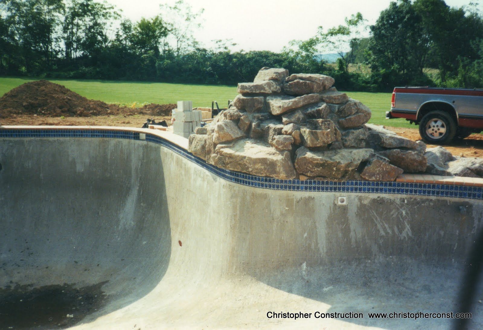 Concrete pool under construction with stacked rocks beside a pickup truck and grassy yard.
