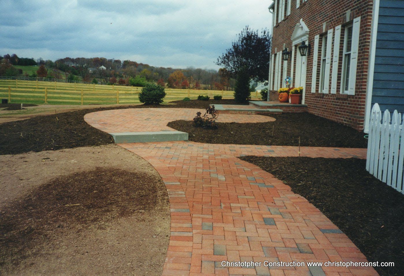Curved brick walkway and landscaped garden beside a brick house overlooking open fields