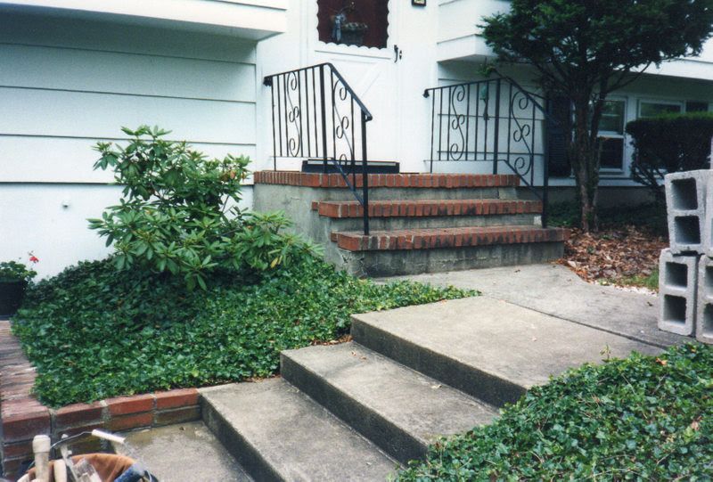 Concrete front steps leading to a porch with black railings, surrounded by green shrubs and ivy.