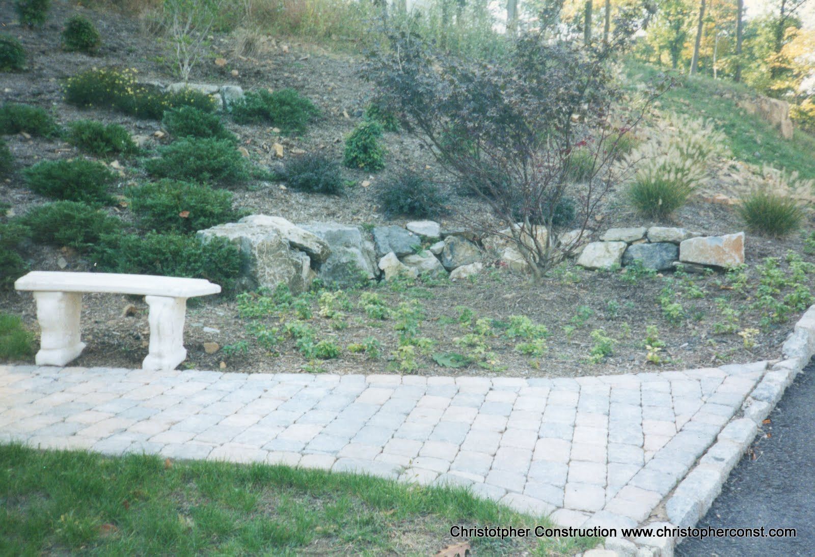 Stone bench beside a paved path in a grassy garden with shrubs and rocks